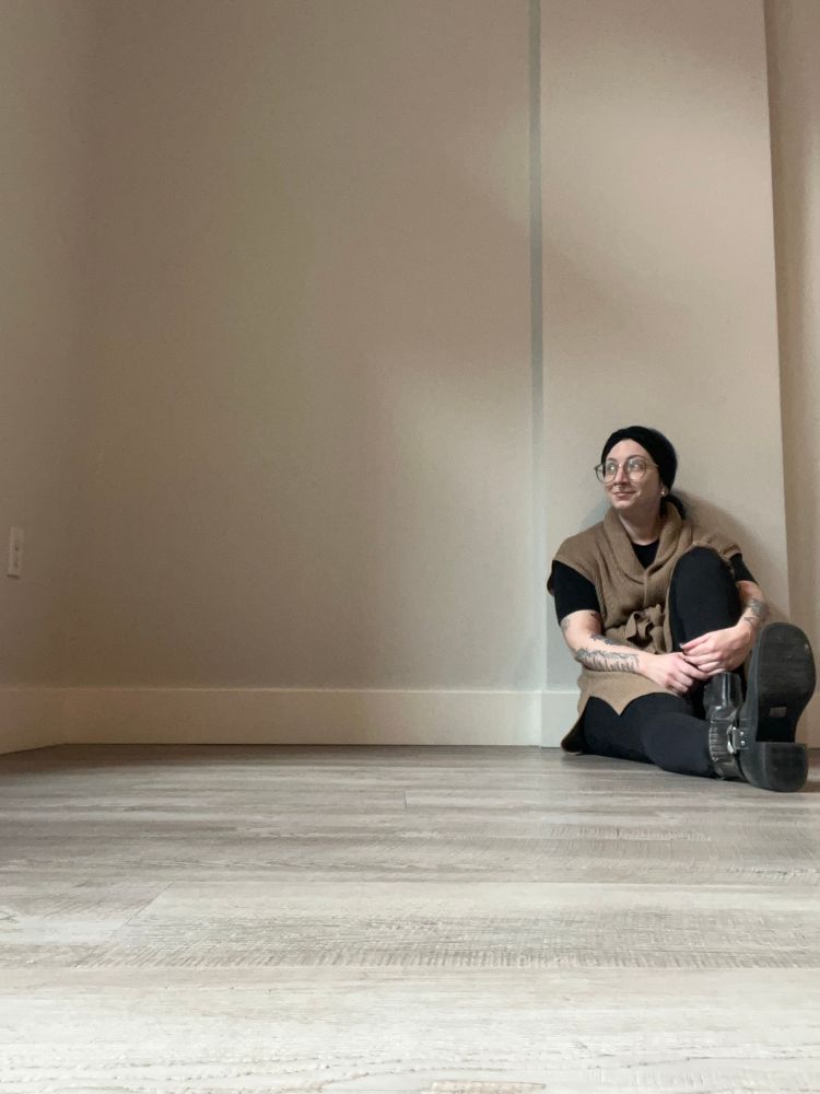 Zoe is sitting up against a blank wall on a wood paneled floor, in an empty room. She is sitting on the right side of the image, looking to the left.

She is wearing glasses, a black headband, a black shirt, black tights, a khaki colored sleeveless cardigan, and black boots. One of her knees is up against her chest, with her arms wrapped around it. She has a soft smile on her face.