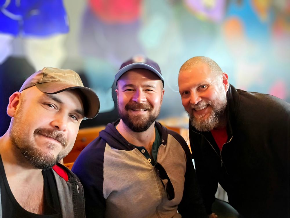 Three bearded men smile for a selfie in a warmly lit indoor space. The man on the left wears a camo cap and an unbuttoned hoodie over a black shirt. The man in the center wears a two-tone hoodie and a navy blue baseball cap, with sunglasses hanging from his collar. The man on the right wears a black zip-up jacket over a red shirt. A colorful mural is visible in the blurred background.