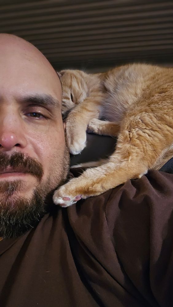 Matt B lies close to his orange cat, who is curled up on his shoulder with a paw resting gently against his face. The cat appears peacefully asleep while Matt B looks toward the camera in a relaxed, intimate moment.