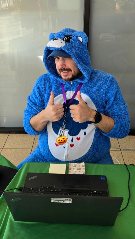 Matt B sits at a table with a laptop, giving two thumbs up while wearing a fuzzy blue onesie designed like a Care Bear with a raincloud and hearts on the belly. He has a work badge and lanyard around his neck, smiling playfully at the camera.