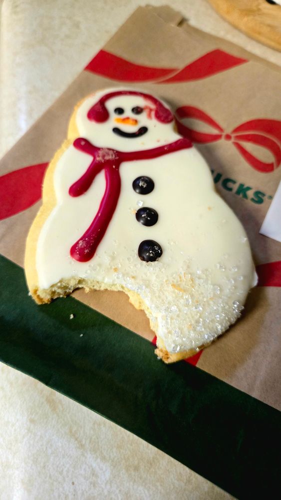 A frosted snowman-shaped cookie with a bite taken out of the bottom sits on a holiday-themed Starbucks bag. The cookie is decorated with white icing, black buttons, red scarf and earmuffs, and sprinkled sugar along the bitten edge.