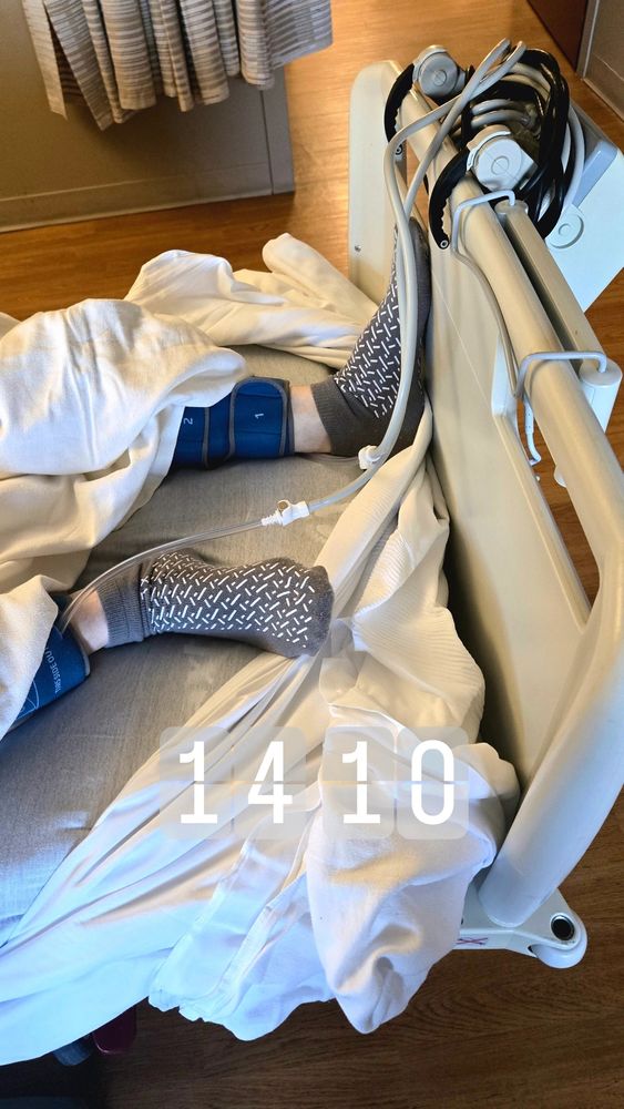 Hospital room scene. A patient lies in a bed with white sheets pulled loosely around their legs. Only the lower legs and feet are visible. They are wearing gray non-slip hospital socks and blue pneumatic compression cuffs. At the side of the bed, several coiled monitor cables hang on the bed rail. The floor is light wood, and a curtain is partially visible in the background. A digital-style overlay on the image reads “1410.”
