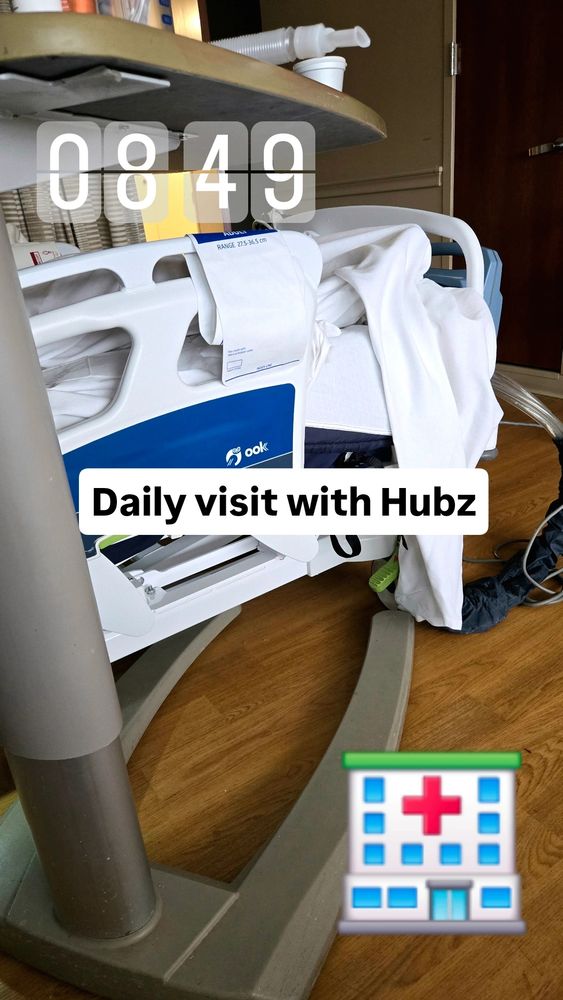 A hospital room scene shows part of a hospital bed with white sheets and a tray table above it. Overlay text reads “0849” and “Daily visit with Hubz,” with a hospital emoji at the bottom right.
