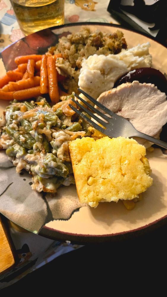 A plate filled with Thanksgiving food, including green bean casserole, glazed carrots, stuffing, mashed potatoes, sliced cranberry sauce, a piece of turkey breast, and a square of cornbread. A fork rests on the plate, and a glass of iced tea sits in the background.
