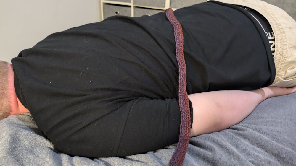 A sleepy young man lying down covered by a crochet blanket, the width of a queen bed and around an inch tall so far.