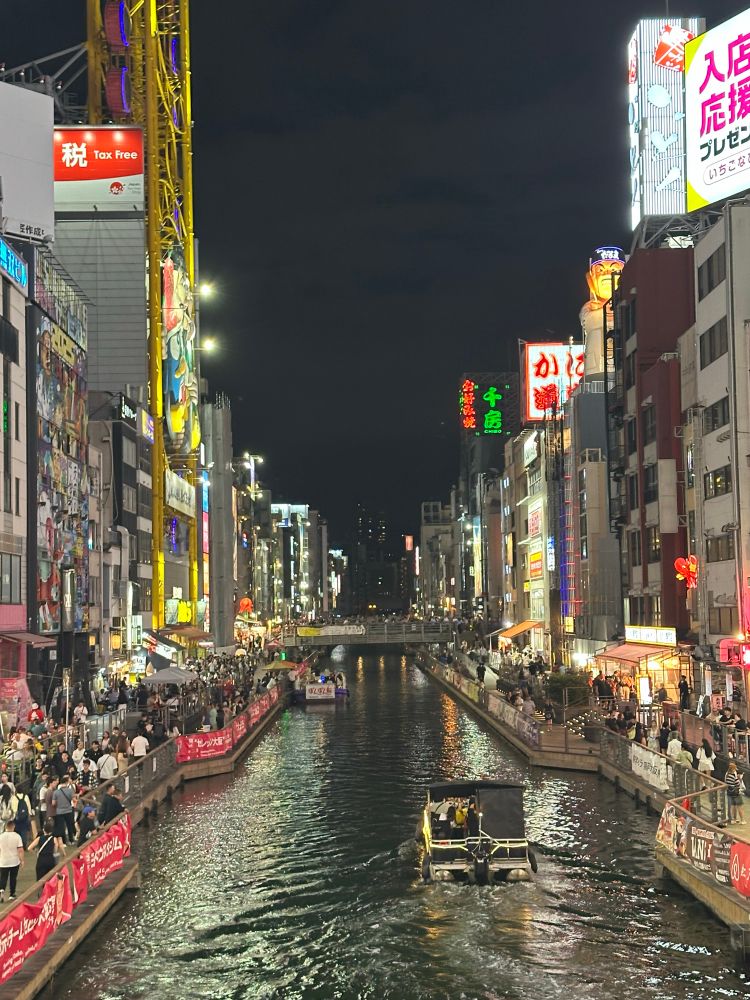 The river running through Dotonbori, with the buildings on the sides lit up with lights and signs. There are big crows on both sides of the river, and a boat is on the water traveling away from the viewer