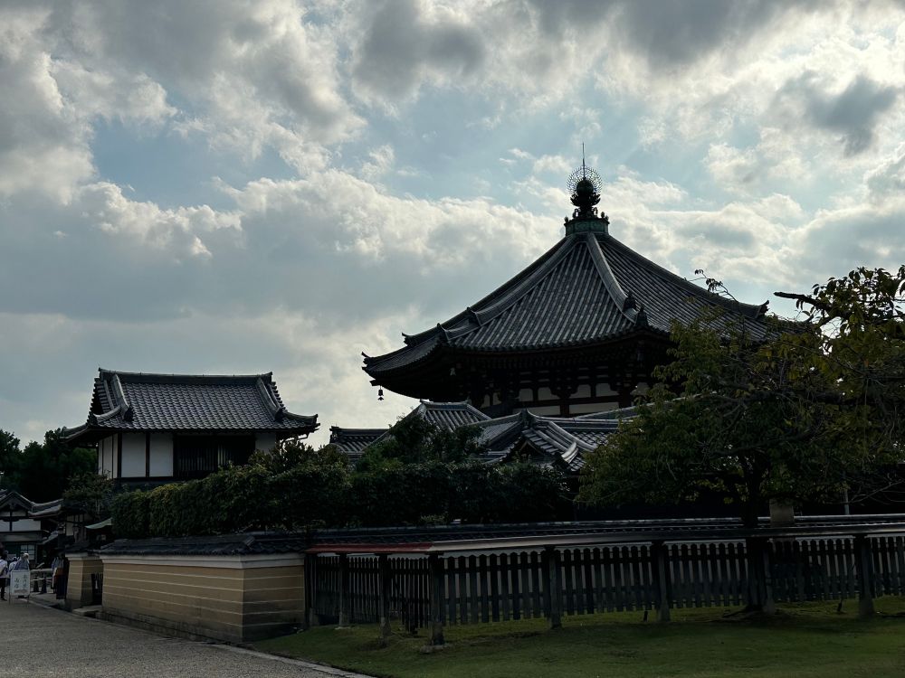 The Southern Round Hall of Kohfukuji in the background, with several buildings and bushes in front, against a cloudy blue sky