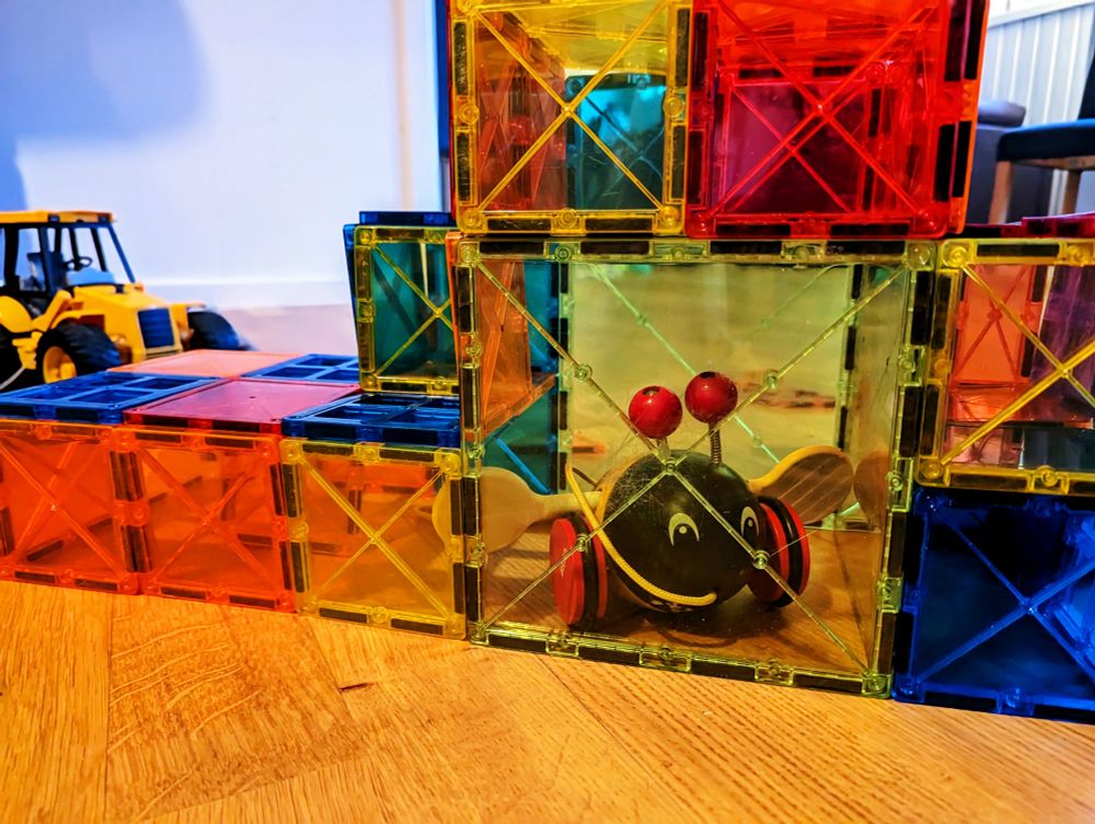 a multi-story structure constructed out of colorful translucent magnetic tiles, containing a classic Swedish pull-toy (a wooden bee on wheels with wings that spin and go brrrr). There's a backhoe in the background