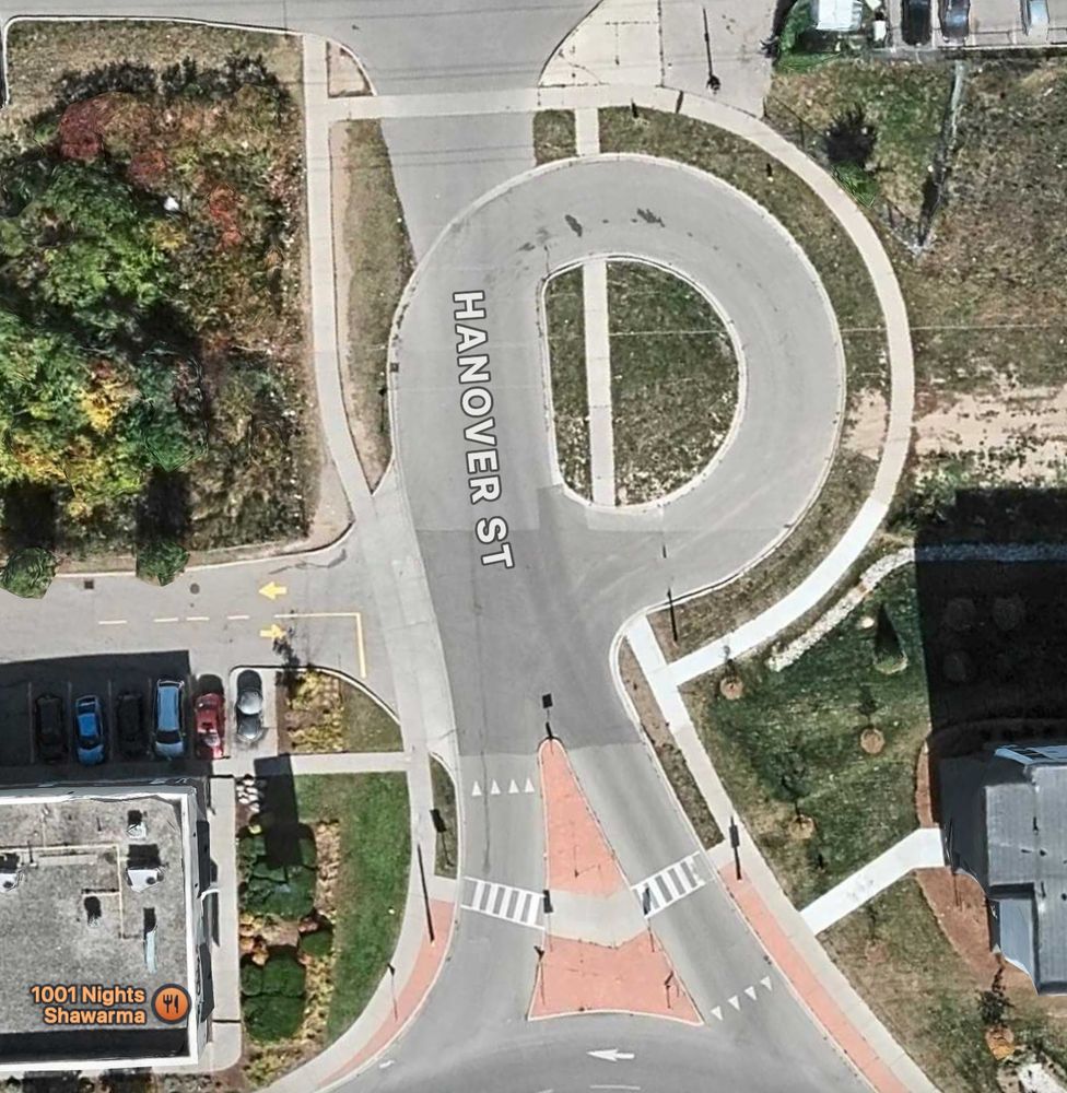 Satellite view of Hanover Street in Kitchener, a little stub off a roundabout that goes past a strip mall entrance, then has a turnaround, and a school driveway off the turnaround. There's a sidewalk going along the turnaround and also over it. It just looks weird