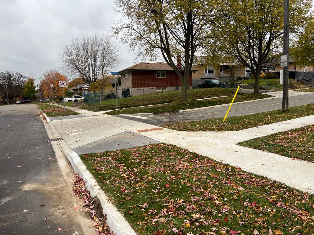 Newly reconstructed low rise residential suburban street meeting a minor collector street, with the road going up to meet a concrete sidewalk that continues across the side street in front of the stop sign