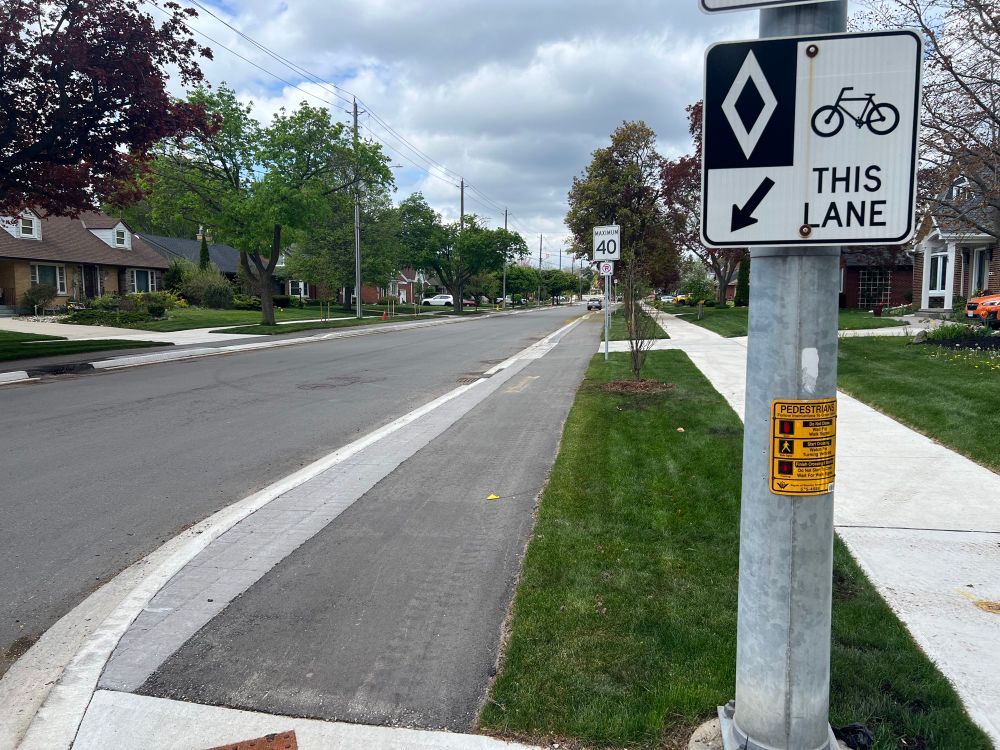 View of newly rebuilt suburban residential street with curb protected asphalt one way bike lane next to the road, a boulevard strip with trees, then sidewalk, and stepped back houses