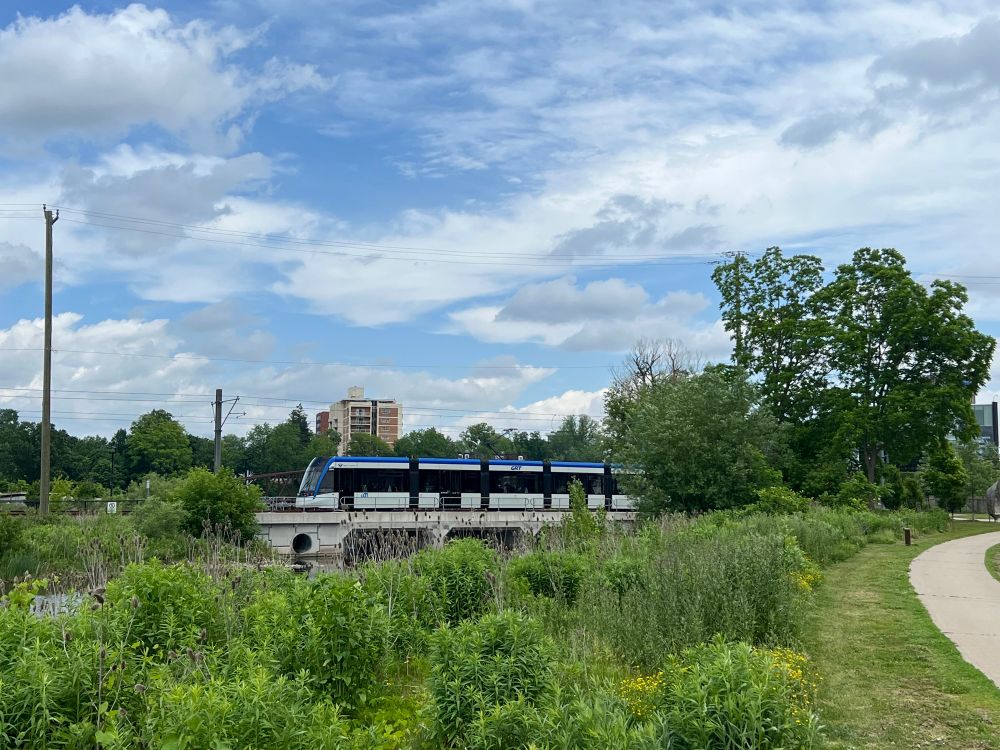 Ion light rail passing over bridge in Waterloo Park under a partly cloudy sky on a summer day