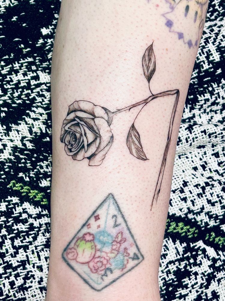 A photo of someone’s arm showing a tattoo that depicts a rose with a broken stem in black ink. There is also an old tattoo of a d4 terrarium in black ink and colour below it. 