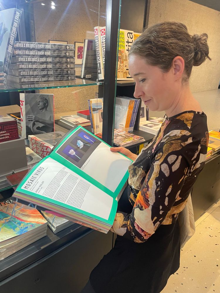 A photo of Rosemary Lee holding the book Kinetismus: 100 Years of Electricity in Art in the gift shop at Kunsthalle Praha