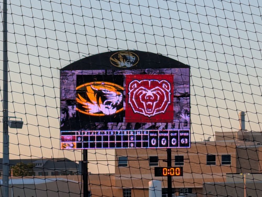 Scoreboard beyond the left center field fence showing no score in the first inning and large logos above it for Missouri and Missouri State. 