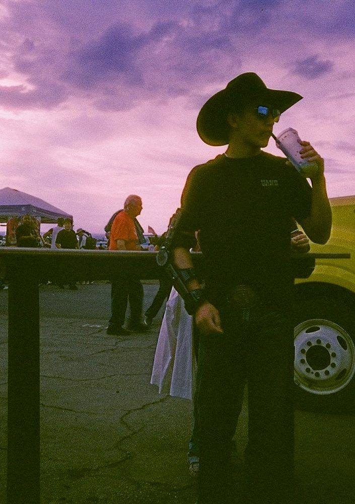 Guy with cowboy hat, sunglasses, a slushie and an arm brace in front of a purplish sunset
