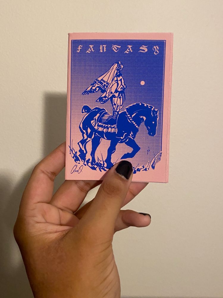 My hand holding a small zine no larger than my palm. The paper is a pale pink with dark blue ink. The cover depicts a woman dressed in loose fabrics standing on the back of a black horse. The sky is a gradient halftone with a small pale moon. The title on the top reads "Fantasy" in wide spaced blackletter.