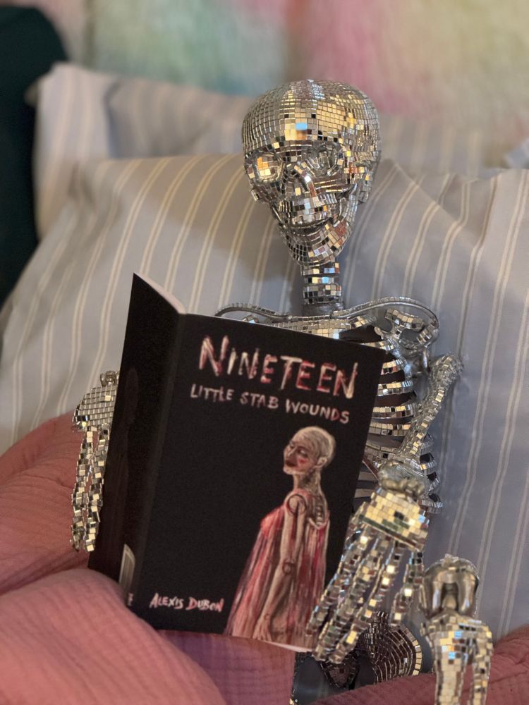 A skeleton made of disco ball mirrors lounges in bed with a book held open in his hands to reveal the cover.

Sparkle Barry is feeling grateful and giving thanks that he doesn’t have any giblets to yank from his disco ball bones because watching those plastic-wrapped organs get pulled out of the turkey was traumatizing! He’s recovering from the gnarly scene with a copy of NINETEEN LITTLE STAB WOUNDS, a chapbook of dark microfiction and flash by Alexis Dubon.