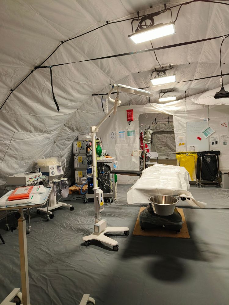Leikkaussali kenttäolosuhteissa valkoisen pressuteltan sisällä. Kuvassa leikkauspöytä ja muuta kirurgista välineistöä. Lattiaa peittää vihreä pressu. 