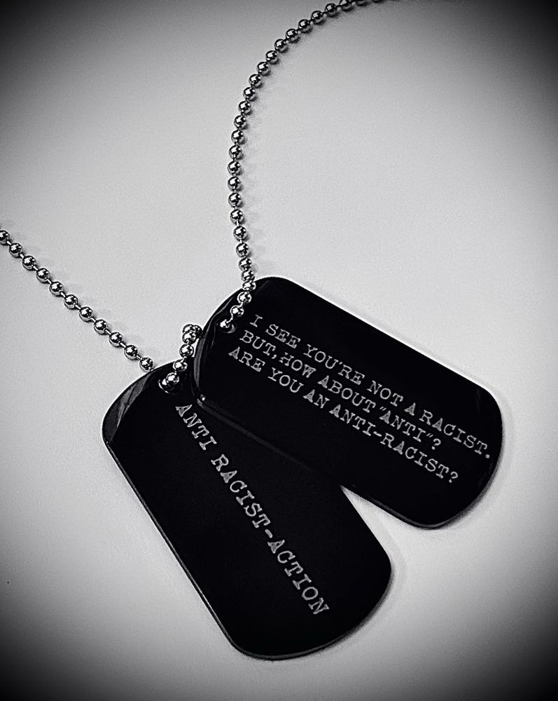 白い背景。黒いDogtag型ペンダント。レーザー刻印で
"I see you're not a racist. But, how about "ANTI"? Are you an Anti-Racist?"(あなたがレイシストであるのかを聞いているのではありません。あなたが反レイシストであるのかを聞いているのです。)と英文が刻まれている。