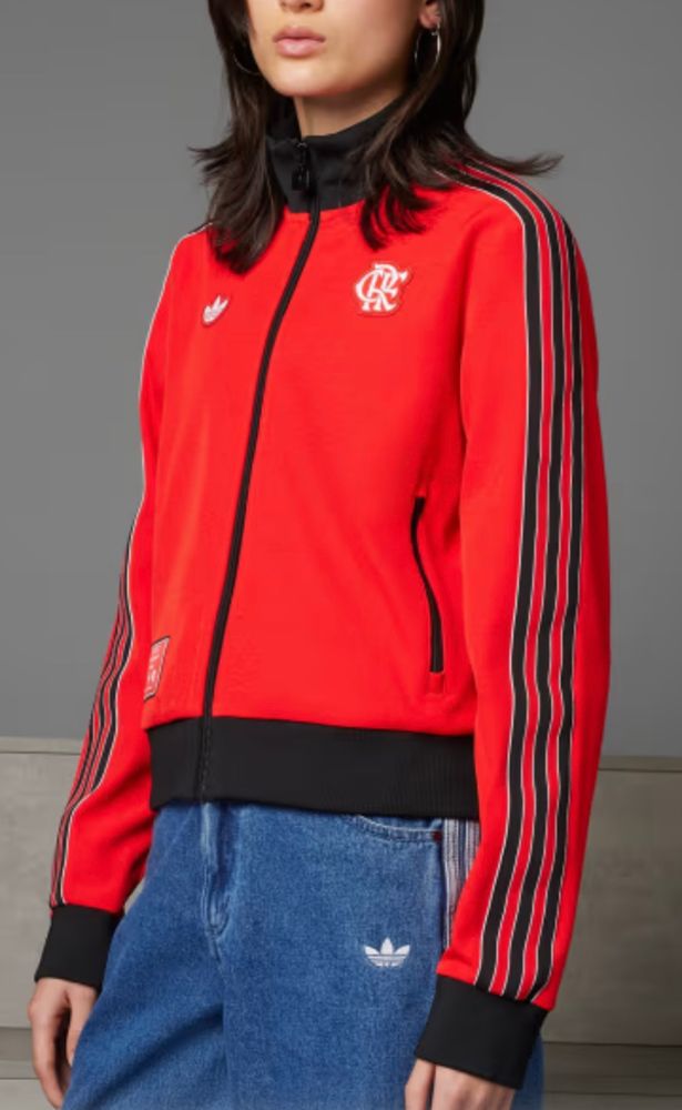 uma mulher de cabelos lisos e castanhos, na altura dos ombros, veste um agasalho esportivo vermelho com detalhes pretos na gola, punhos e cintura, nas laterais dos braços há 3 listras pretas finas, na altura do peito ao lado esquerdo está bordado, em branco, o escudo do clube de regatas do flamengo, do lado direito a logo da adidas, composto por três folhas, também em branco.