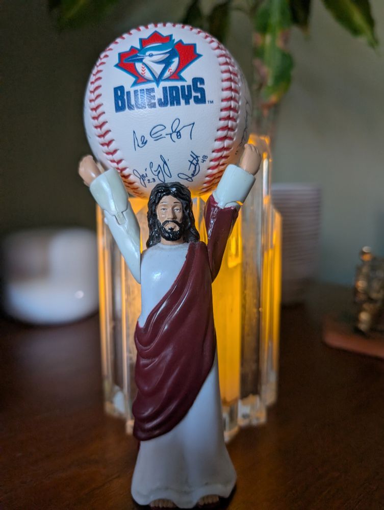 A Jesus action figure with arms raised, holding up a Blue Jays baseball, and standing in front of a lit candle.