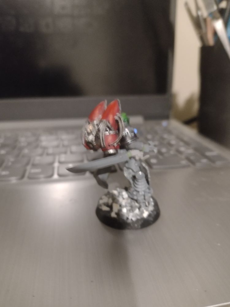 A kitbashed Raven Guard space marine character, with a sword and plasma pistol and a jetpack. The same model as previous, but from the back
The bits are visibly of different origins, some being painted and others bare plastic. He is kicking off of a technological looking piece of rubble