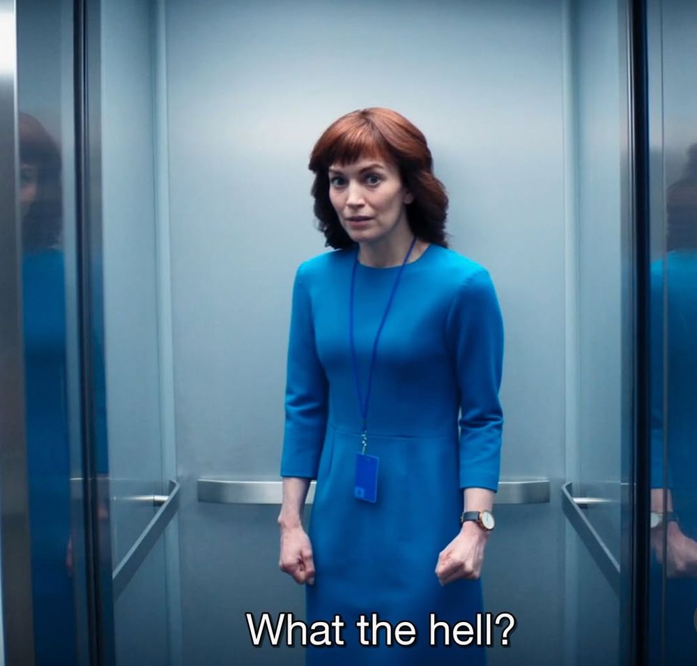 Hellie from Severance. She is a white woman with red hair, bangs straight across her face. She looks shocked. She is wearing a blue dress that covers most of her body, and a blue lanyard with a blue keycard. She has a watch on with a grey band on her right wrist. She is in an elevator, and the tone of the picture is fairly monochromatic. The text at the bottom in white reads "what the hell?"
