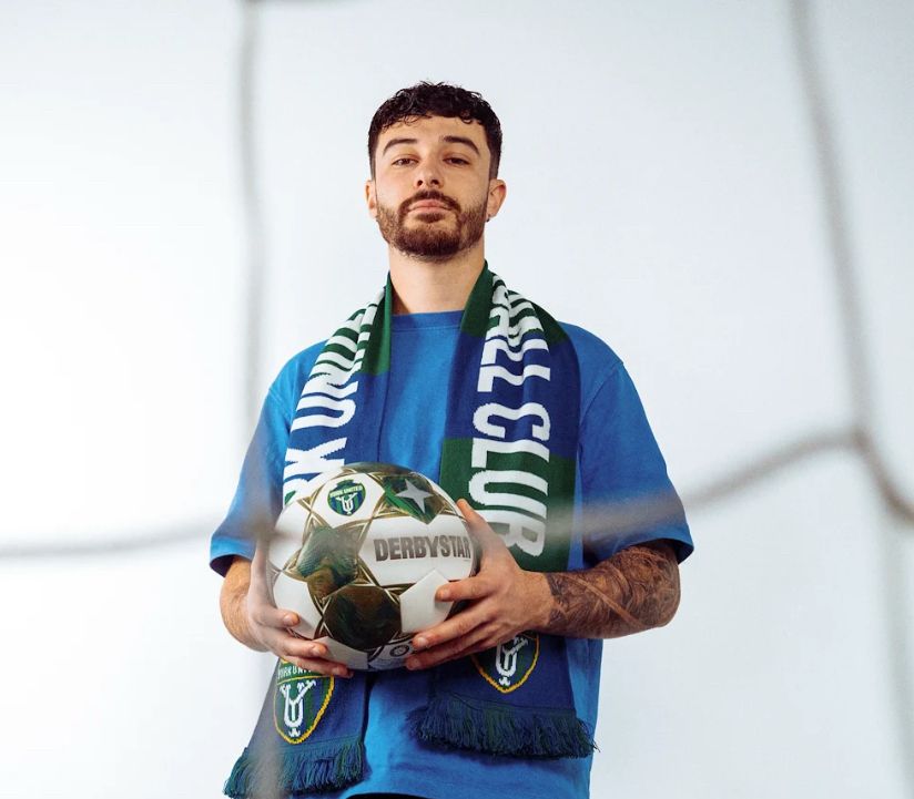 Riley Ferrazzo wearing a York United scarf and holding a soccer ball, pictured from behind the mesh of a soccer net