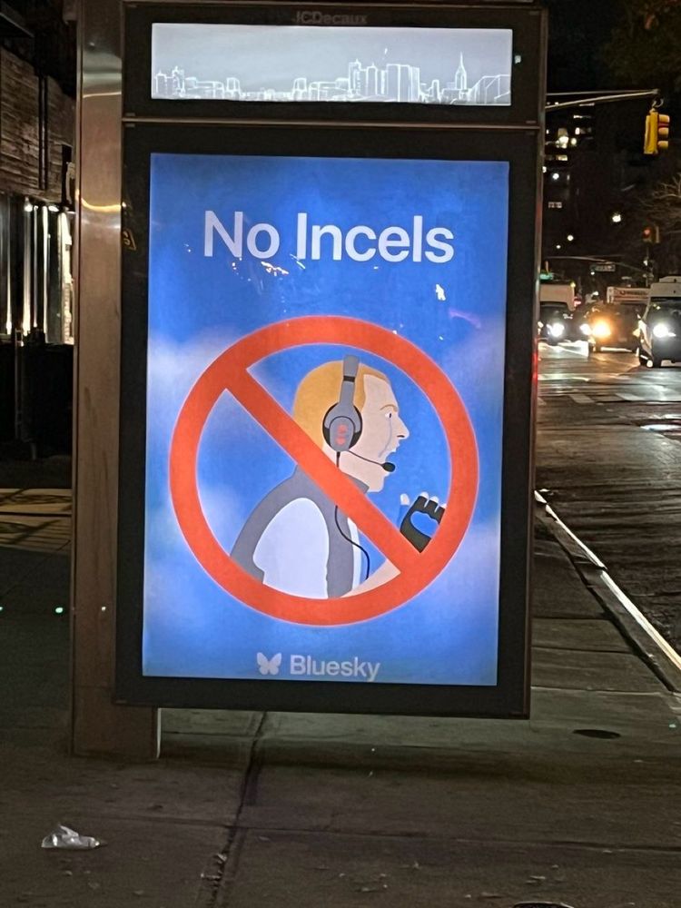 
An image of a bus stop advertisement with the text “No Incels” written in bold white letters against a blue background. Below the text, there is an illustration of a person wearing a headset and holding a microphone, crossed out by a red circle with a diagonal line, indicating prohibition. At the bottom, the logo and name “Bluesky” are visible. The setting appears to be an urban street at night, with cars and streetlights in the background.