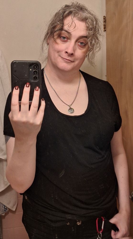 A woman with gray brown hair wearing a black tshirt, pentacle necklace, eye shadow and a smirk looking exhausted having just finished the 4th consecutive day of driving across the country.