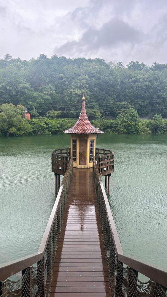 A small bathing house with a pointed roof sits at the end of a narrow boardwalk above a blue lake. There are green bushes at the end of the lake behind the bathing house.