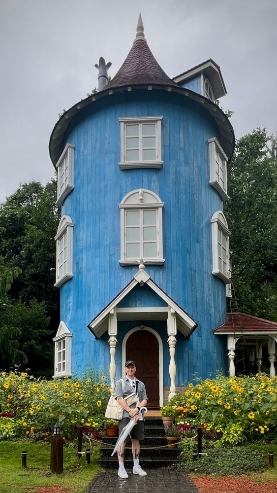 A blue cylindrical three story house surrounded by flowers punctuates a greenery filled scene. A man stands in front of the house doorway with an umbrella in hand.