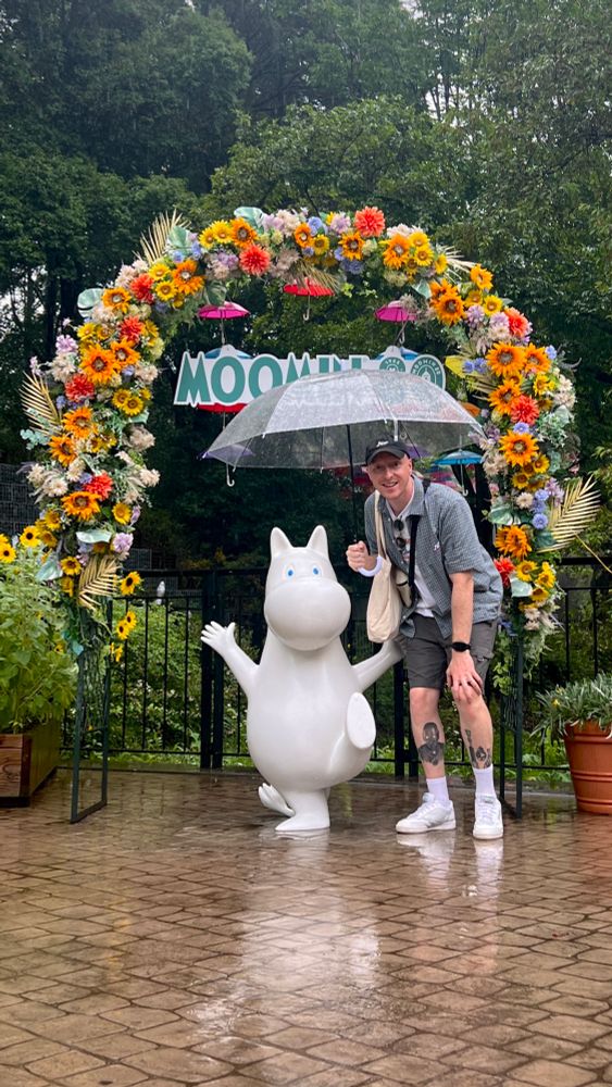 A man stands beneath a floral arch next to a Moomin. It is raining and he is holding an umbrella over their heads.