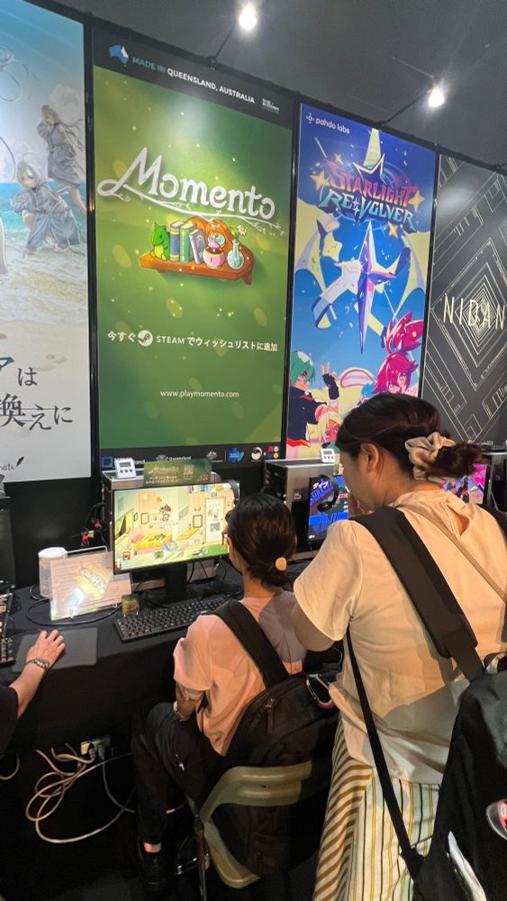 A woman watches as her friend sits at a computer monitor playing the upcoming video game Momento.