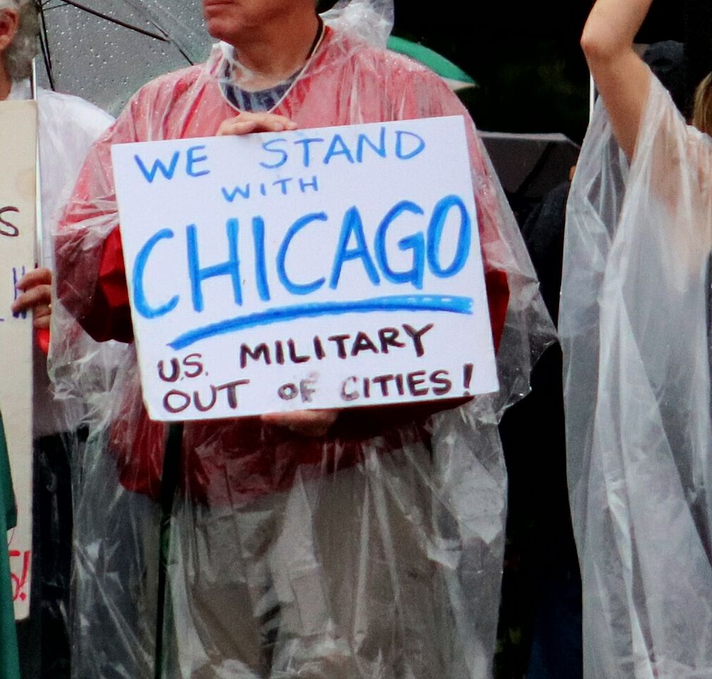 Older white man at No Kings rally in Edwardsville, IL holds sign that says "we stand with Chicago / U.S. military out of cities!"