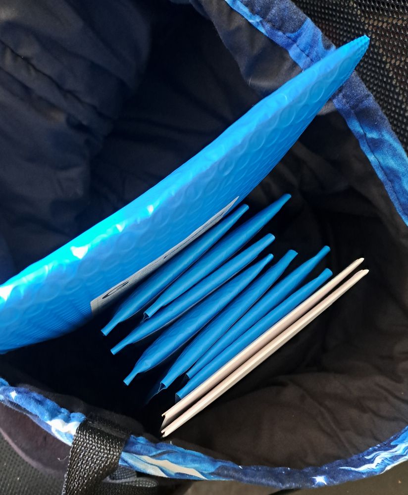 A photograph of 10 small packages in a blue tote bag. One is in a very large blue bubble mailer, placed at the back of the stack. In front of it are 7 smaller blue mailers, and in front of those are 2 white cardboard envelopes. They contain merch orders from my store.