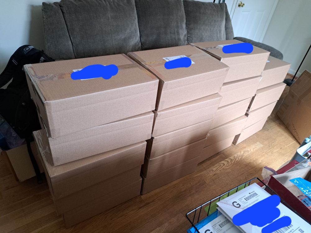A photograph of 19 cardboard boxes stacked in front of a grey couch in a living room. They are packages containing ita bag orders from my store.