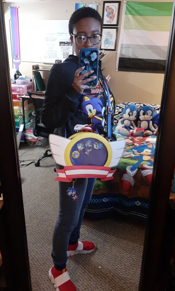 Photograph of a black woman taking a selfie in a bedroom. She's wearing a Sonic Adventure 2 jacket, and is wearing an ita bag shaped like the emblems on title screens of various Sonic the Hedgehog games. There is a bed behind her with Sonic bedsheets and plushies on it. There is also a desk space with boards and artwork on the walls behind her.