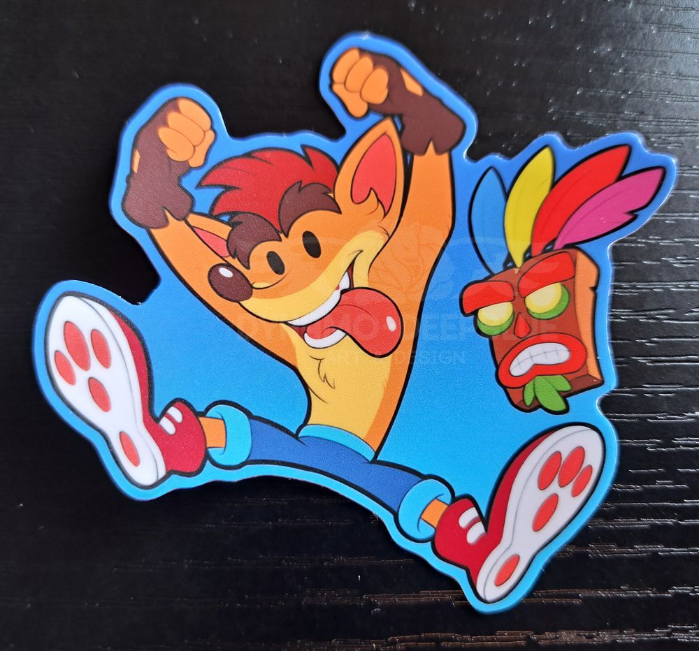 A photograph of a Crash Bandicoot-themed sticker with art by Dynamo-Deepblue. Crash is shown jumping excitedly with his tongue hanging out and Aku Aku by his side, with a blue border with a lighter blue gradient on the lower half. The sticker is placed on a black wooden desk.