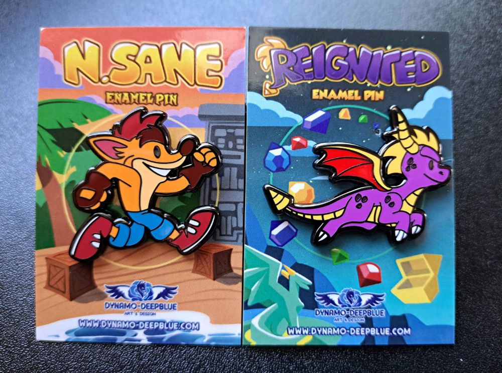 A photograph of two enamel pins depicting a chibi Crash Bandicoot and a chibi Spyro the dragon, both with themed backing cards, placed side by side, on a black surface.