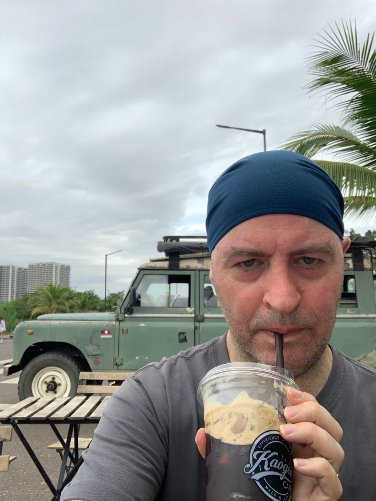 Me looking very red drinking an iced coffee with a beat-up Landrover in the background.