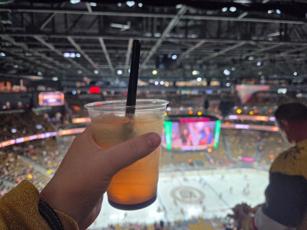 A hand holding a golden cocktail in a plastic cup with T Mobile arena in the background 