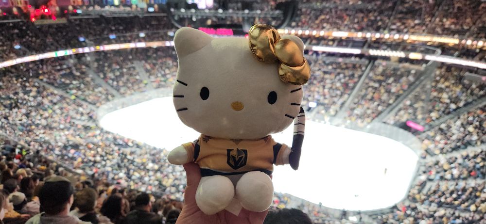 A VGK hello kitty plush being held up with an NHL arena in the background 