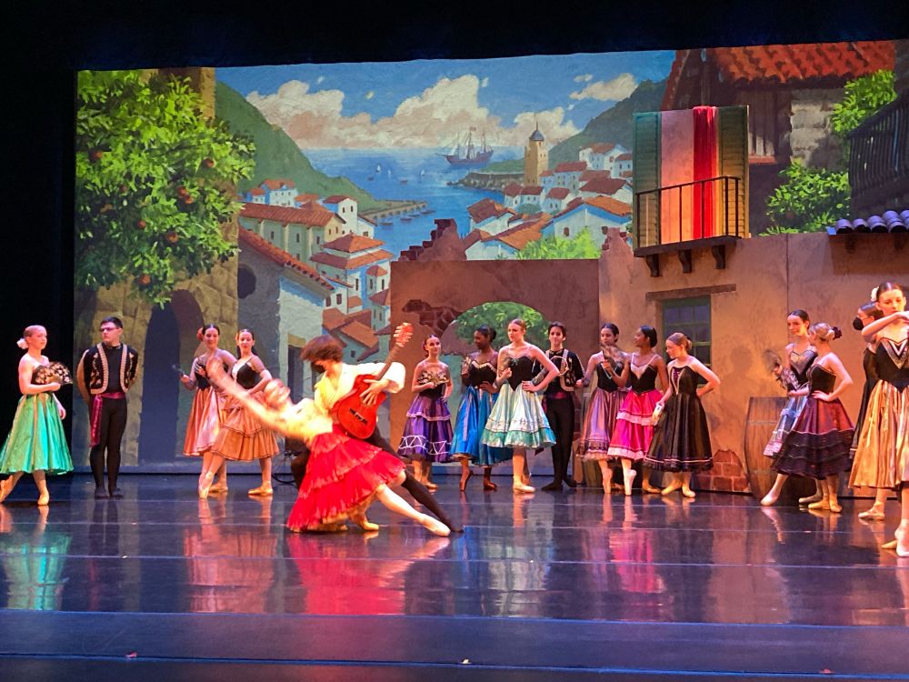 Ballet dancers on stage in Spanish outfits, backdrop of a Spanish seaside town.