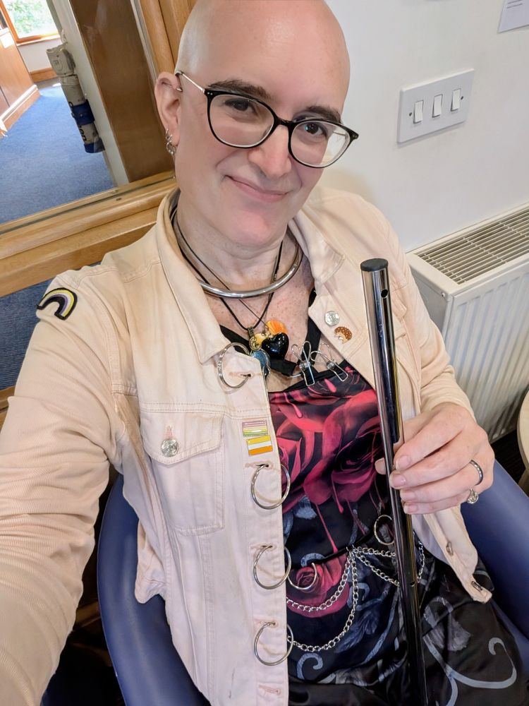 A selfie of Jaz sat on a chair at Parkside Hospital. Fae is carrying a spreader bar minus it's split rings, with the rings looped through the button holes of faer faded peach pink denim jacket.