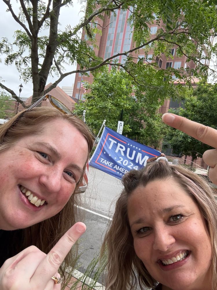 Two ladies holding up their middle fingers to a Trump flag behind them.