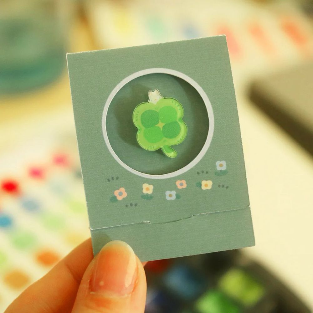 A pic of a hand holding a folded backing card that contains a 4-leaf clover-shaped acrylic pin