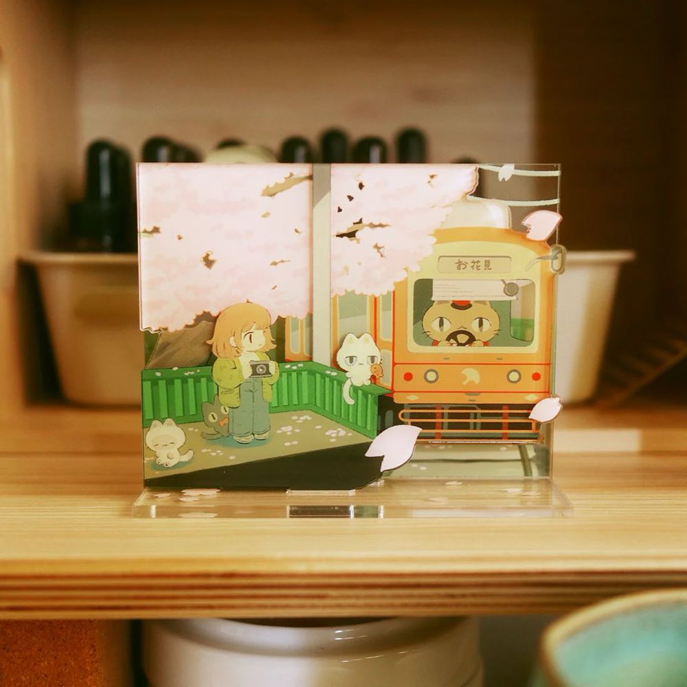 Photo of a hanami train themed acrylic standee featuring a red train going to the cherry blossom fields, while a cat conductor drives it, and a girl with 3 quirky cats waits to board the train, camera in hand