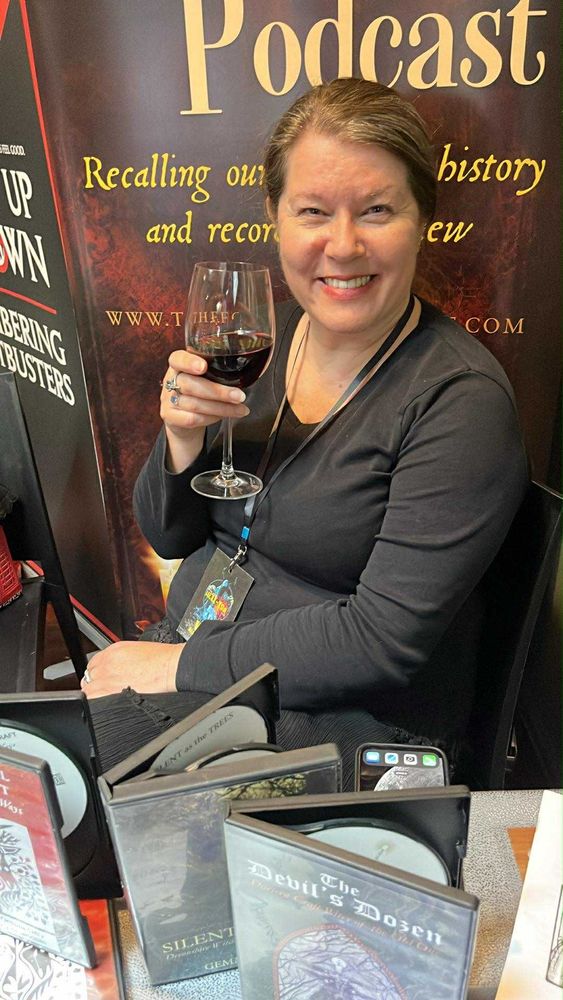Shot of woman sitting behind a table, glass of red wine in hand. In front of her are some audiobook CDs for sale. The woman is happy because she has fulfilled her obligations for the weekend and is now free to relax. Yes, the woman is me. :) 